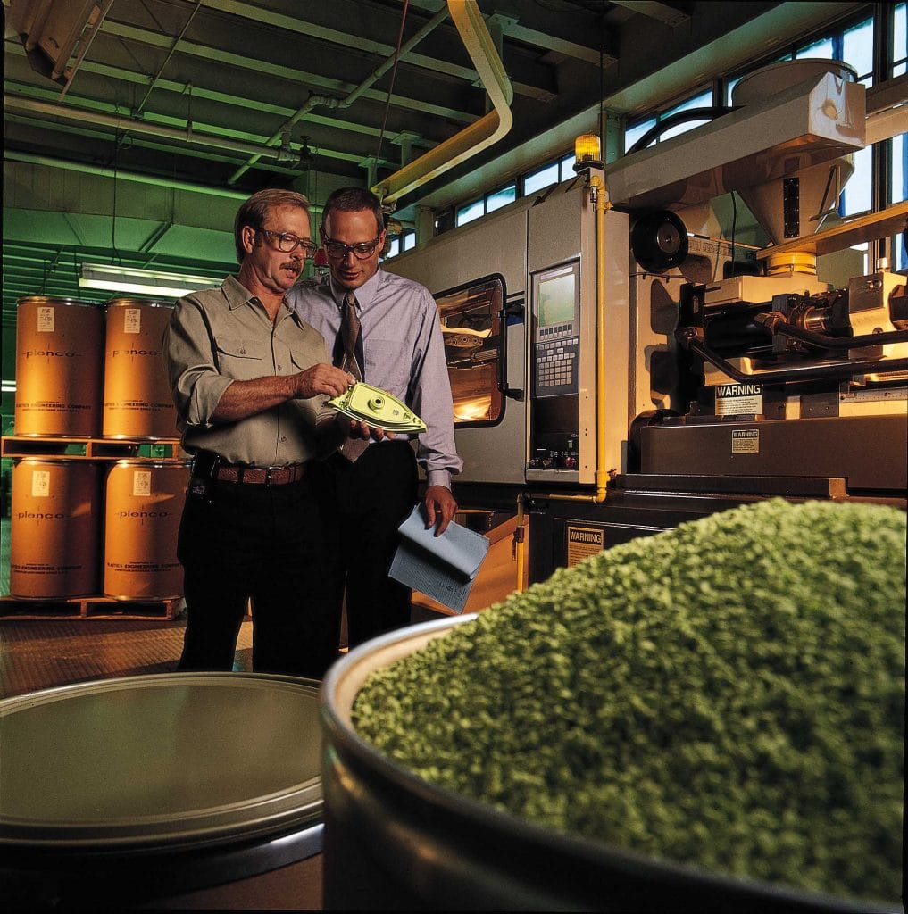 PLENCO professionals inspecting a thermoset molded iron component next to an injection molding machine and barrels of raw material.