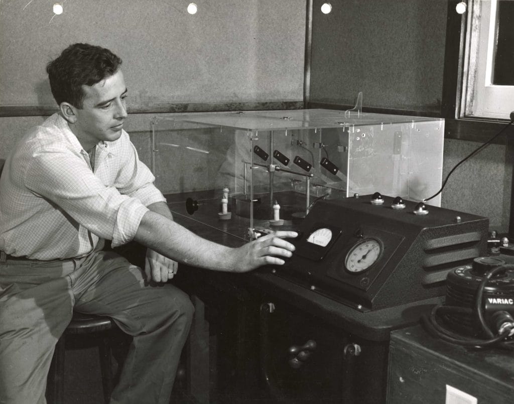A vintage black-and-white photo of a PLENCO technician operating a precision testing apparatus in a laboratory setting.