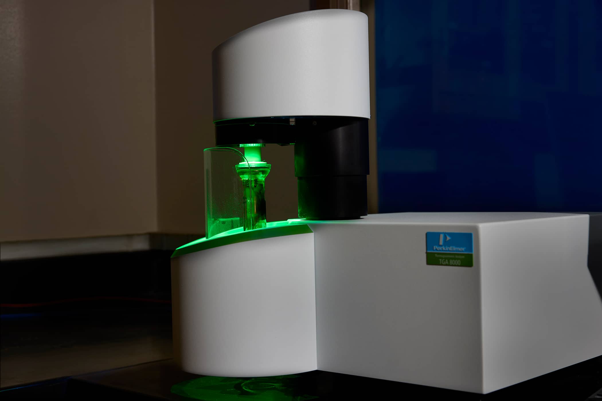 A PerkinElmer TGA 8000 thermogravimetric analyzer in a PLENCO laboratory, illuminated with a green light during material testing.