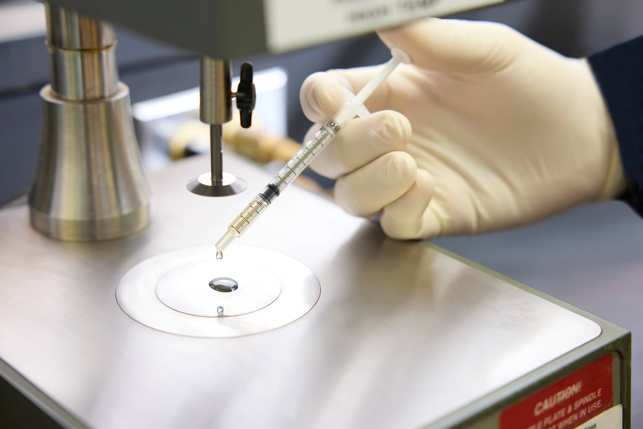 A laboratory technician in a gloved hand using a syringe to carefully place a liquid sample onto a precision testing plate.