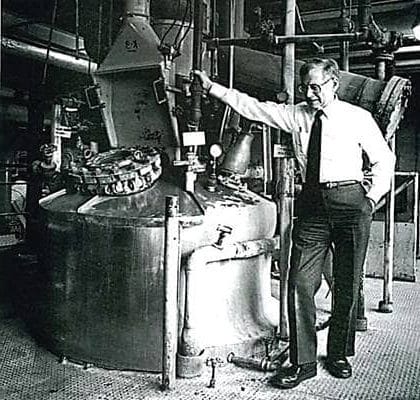 A vintage black-and-white photograph of a PLENCO engineer standing next to a large industrial reactor vessel.