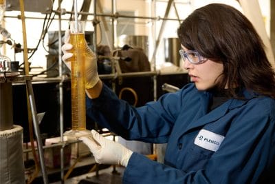 A PLENCO chemical engineer in a blue lab coat and safety glasses inspecting a liquid sample in a graduated cylinder.